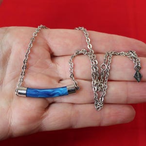 May include: A silver-toned necklace featuring a blue and white marbled tube pendant. The pendant is encased in silver metal. The chain is a delicate link style, and the necklace is displayed against a red background.