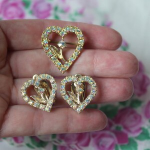 May include: A set of three gold-tone heart-shaped brooches with clear iridescent rhinestones. The brooches feature a floral design in the centre.