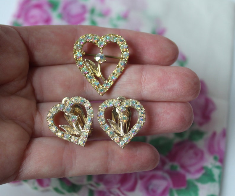 May include: A set of three gold-tone heart-shaped brooches with clear iridescent rhinestones. The brooches feature a floral design in the centre.