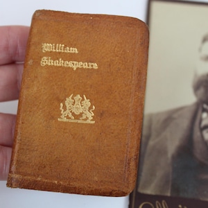Peut inclure: Un petit livre en cuir marron avec des lettres dor&eacute;es "William Shakespeare" et un embl&egrave;me dor&eacute;. Un portrait s&eacute;pia d'un homme barbu est en arri&egrave;re-plan.