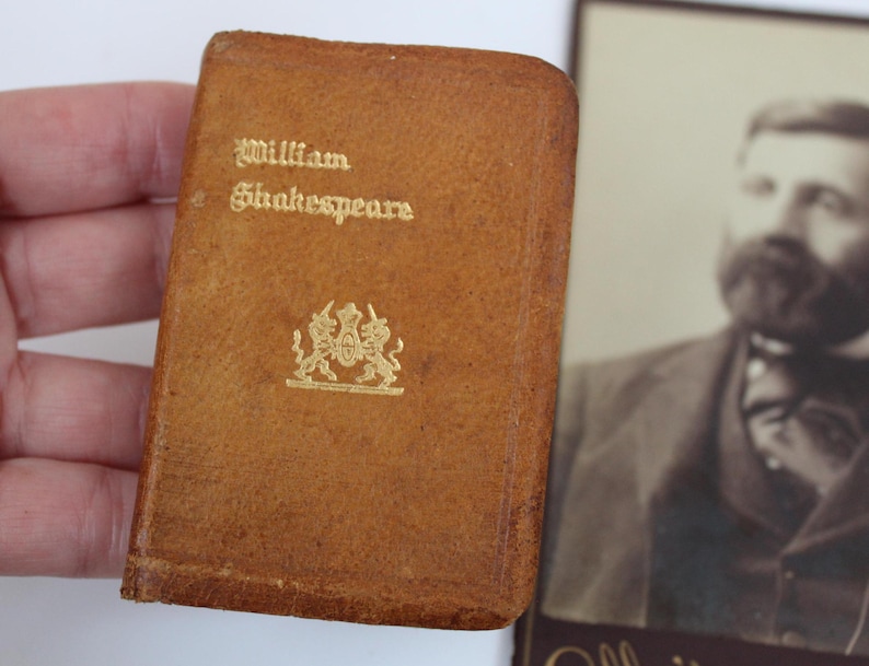 Peut inclure: Un petit livre en cuir marron avec des lettres dor&eacute;es "William Shakespeare" et un embl&egrave;me dor&eacute;. Un portrait s&eacute;pia d'un homme barbu est en arri&egrave;re-plan.