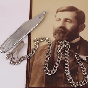 May include: A silver pocket knife with a chain and clasp, next to a vintage photograph of a man with a beard. The knife has a textured design. The chain is made of small, interlocking links.