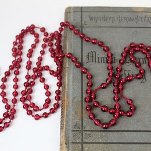 May include: A long strand of deep red, translucent spherical beads strung together on a thin cord. The necklace is draped across a weathered, gray-toned hardcover book titled Whitney's German Texts with the word Minna von Barnhelm visible.