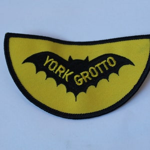 May include: A yellow patch with a black bat silhouette and the words "YORK GROTTO" embroidered in black. The patch has a black border and a curved shape.