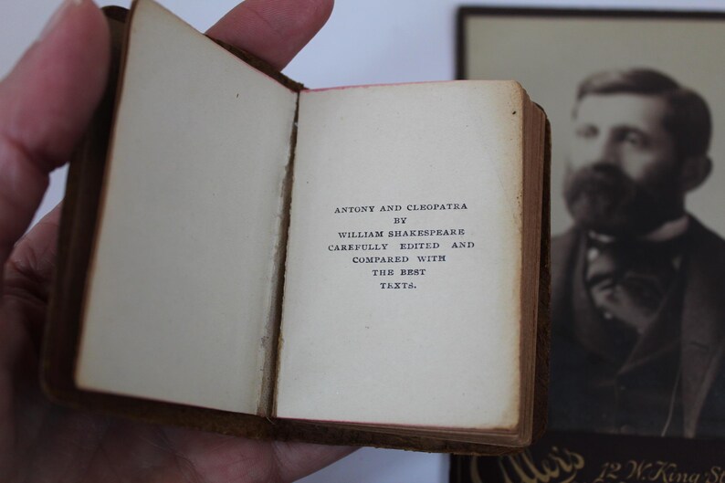Peut inclure: Un petit livre ouvert avec le titre "Antony and Cleopatra" de William Shakespeare. Le texte est imprim&eacute; &agrave; l'encre noire sur des pages blanc cass&eacute;. Le livre est tenu ouvert, r&eacute;v&eacute;lant le texte, avec une couverture en cuir marron.