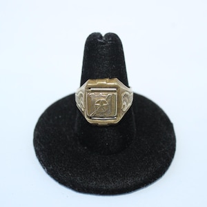 May include: A gold-toned ring with a rectangular face featuring a star and shield design. The ring is set on a black display stand against a white background. The ring's band has etched details on the sides.