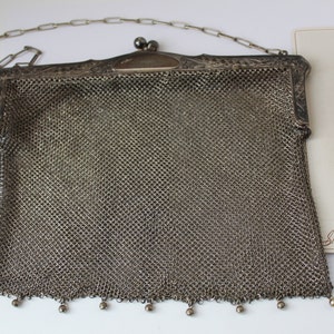 May include: A silver mesh purse with a chain strap and a decorative clasp. The purse has a beaded fringe at the bottom.