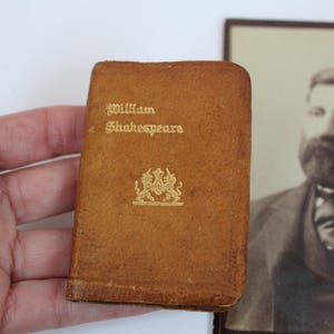 Peut inclure: Un petit livre ancien avec une couverture en cuir marron. La couverture porte l'inscription dor&eacute;e &laquo; William Shakespeare &raquo; et un embl&egrave;me dor&eacute;. Un portrait en noir et blanc d'un homme est en arri&egrave;re-plan.