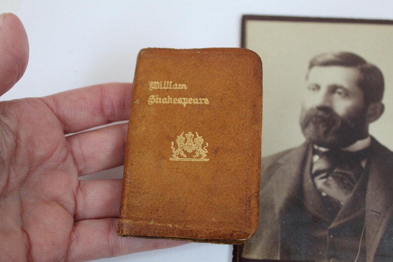 Peut inclure: Un petit livre ancien avec une couverture en cuir marron. La couverture porte l'inscription dor&eacute;e &laquo; William Shakespeare &raquo; et un embl&egrave;me dor&eacute;. Un portrait en noir et blanc d'un homme est en arri&egrave;re-plan.