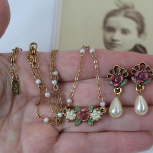 May include: A gold-toned necklace and earrings set featuring a floral design. The necklace has a pendant with red roses, white pearls, and green leaves. The earrings have a similar design with teardrop-shaped pearls. The necklace has a tag that reads "1928".