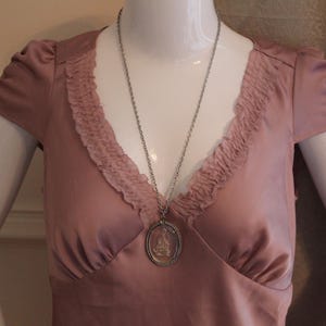 May include: A dusty rose-colored dress with a V-neckline and ruffled trim. A silver-toned necklace with a pendant featuring a detailed design hangs on the dress. The dress has short sleeves and is displayed on a mannequin.