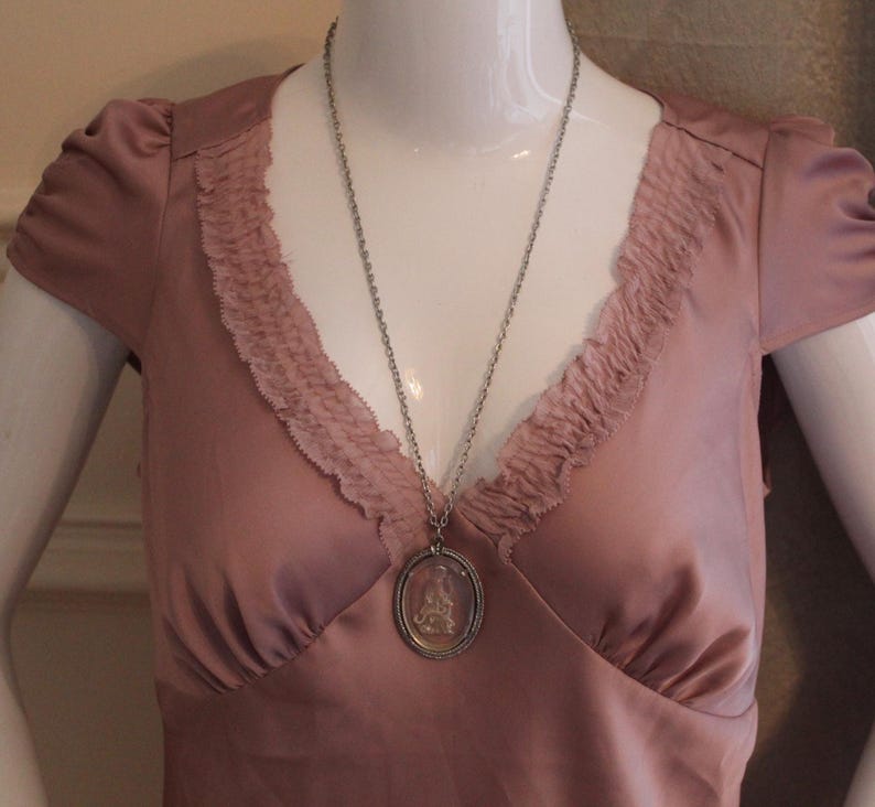 May include: A dusty rose-colored dress with a V-neckline and ruffled trim. A silver-toned necklace with a pendant featuring a detailed design hangs on the dress. The dress has short sleeves and is displayed on a mannequin.