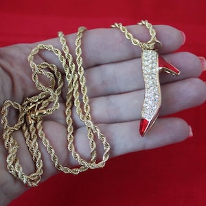 Puede incluir: Un collar de cadena de cuerda dorada con un colgante en forma de zapato rojo de tacón alto incrustado con diamantes de imitación transparentes.