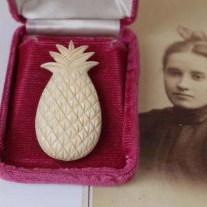 May include: A cream-colored pineapple brooch with a carved diamond-patterned surface and pointed leaves at the top. The brooch is displayed inside an open, rectangular jewelry box lined with deep magenta velvet and white satin fabric.