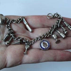 Pode incluir: Um bracelete de prata com uma variedade de pingentes, incluindo um futebol, uma faca, uma garrafa, um copo, um dedal, um alfinete e um pingente redondo com o texto "Mohawk Trail Mass".