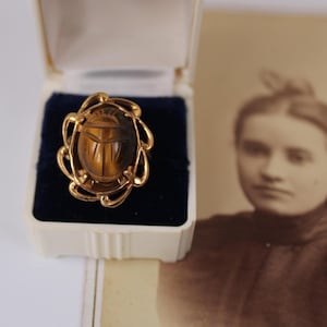 May include: A gold-toned ring with a carved scarab beetle design, set in a decorative frame. The ring is displayed in a white and blue velvet jewelry box. A sepia-toned photograph is in the background.