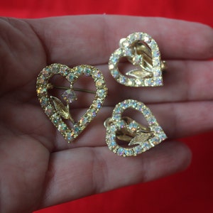 May include: A set of three gold-tone heart-shaped earrings with clear iridescent rhinestones and a floral design.