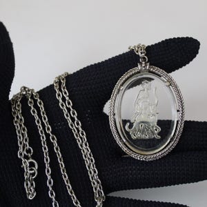 May include: Silver-toned oval pendant necklace featuring a detailed engraved design within a clear, oval-shaped center. The pendant is suspended from a delicate chain. The necklace is displayed against a black background.