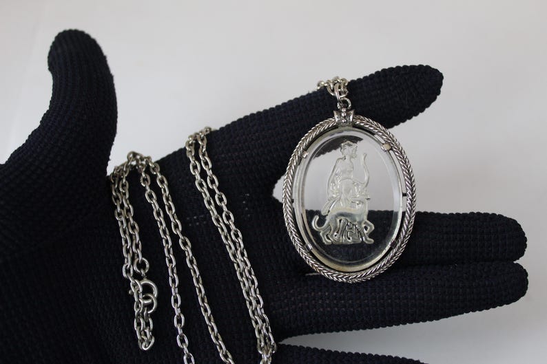 May include: Silver-toned oval pendant necklace featuring a detailed engraved design within a clear, oval-shaped center. The pendant is suspended from a delicate chain. The necklace is displayed against a black background.