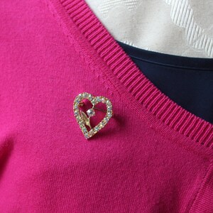 May include: A gold heart-shaped brooch with clear rhinestones. The brooch is pinned to a pink sweater.