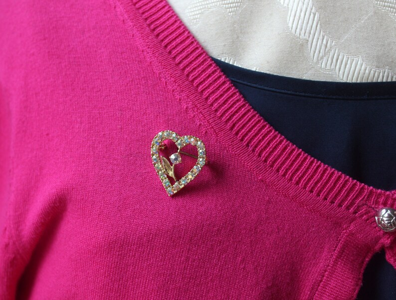 May include: A gold heart-shaped brooch with clear rhinestones. The brooch is pinned to a pink sweater.