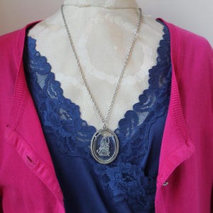 May include: A silver-toned pendant necklace with an oval-shaped, clear glass pendant featuring a detailed figure. The necklace hangs over a navy blue top with lace trim, and a bright pink cardigan.
