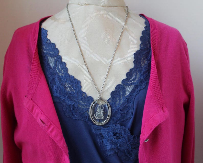 May include: A silver-toned pendant necklace with an oval-shaped, clear glass pendant featuring a detailed figure. The necklace hangs over a navy blue top with lace trim, and a bright pink cardigan.