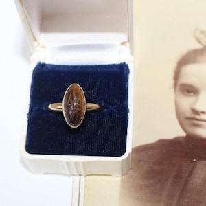 May include: A gold ring featuring an oval-shaped brown stone with an engraved scarab design, presented in a white jewelry box with a dark blue velvet interior lining, positioned next to a vintage sepia-toned photograph of a person.