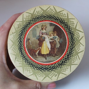 May include: A round, cream-colored tin with a black and white geometric design. The center of the tin features a colorful illustration of a woman and two children in a garden setting.