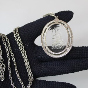 May include: Silver-toned pendant necklace featuring an oval pendant with a detailed relief of a classical figure. The pendant is suspended from a delicate chain. The necklace is held against a dark background, highlighting the silver color and intricate design.