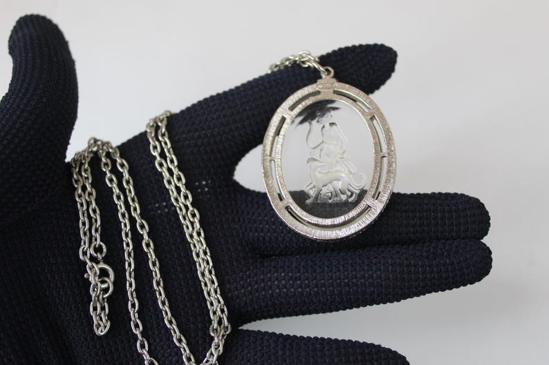 May include: Silver-toned pendant necklace featuring an oval pendant with a detailed relief of a classical figure. The pendant is suspended from a delicate chain. The necklace is held against a dark background, highlighting the silver color and intricate design.