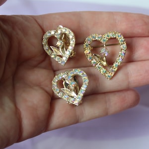 May include: A set of three gold-tone heart-shaped earrings with clear iridescent rhinestones. The earrings feature a delicate floral design with a dangling rhinestone in the centre.