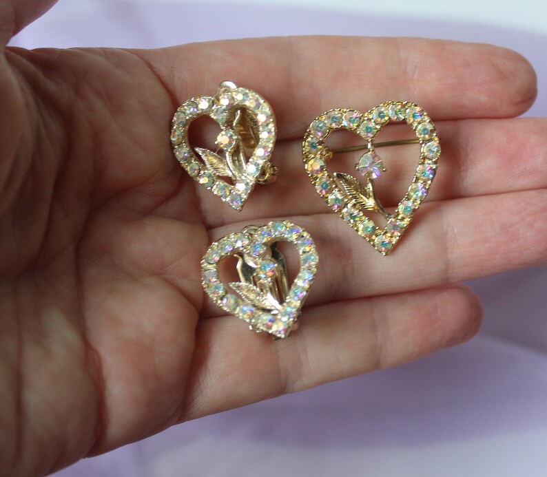 May include: A set of three gold-tone heart-shaped earrings with clear iridescent rhinestones. The earrings feature a delicate floral design with a dangling rhinestone in the centre.