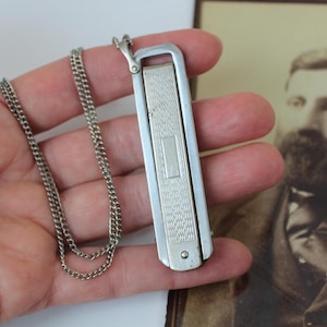 May include: A silver-colored, rectangular pendant with a patterned design and a small chain. The pendant is held in a hand, with a vintage photograph in the background. The chain is approximately 18 inches long.