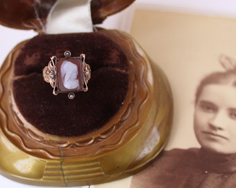 May include: A vintage cameo ring with a rectangular, reddish-brown stone featuring a white profile. The ring is set in a gold-toned metal with ornate detailing and small pearl accents. The ring is displayed in a velvet-lined, antique jewelry box, with a sepia-toned photograph in the background.