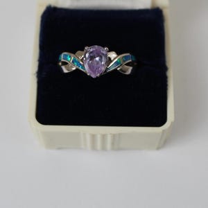 May include: A silver-toned ring featuring a central pear-shaped lavender gemstone. The band is accented with inlaid blue iridescent stone segments that curve along the sides, creating a split-shank design, presented in a dark blue velvet box.