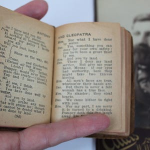 Peut inclure: Un petit livre ancien, aux pages jaunies par le temps, est ouvert, r&eacute;v&eacute;lant du texte imprim&eacute; d'une pi&egrave;ce de th&eacute;&acirc;tre. Le livre a une couverture marron fonc&eacute; et le texte est noir. Un portrait flou d'un homme est en arri&egrave;re-plan.