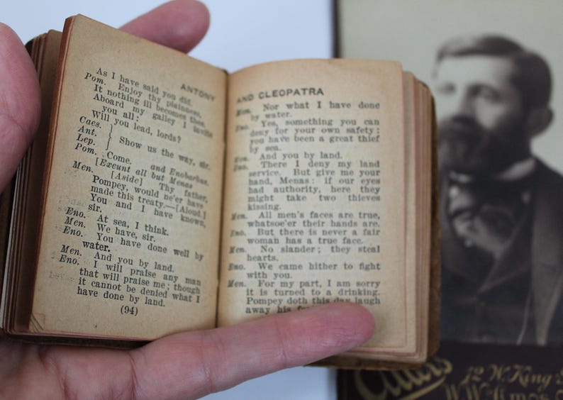 Peut inclure: Un petit livre ancien, aux pages jaunies par le temps, est ouvert, r&eacute;v&eacute;lant du texte imprim&eacute; d'une pi&egrave;ce de th&eacute;&acirc;tre. Le livre a une couverture marron fonc&eacute; et le texte est noir. Un portrait flou d'un homme est en arri&egrave;re-plan.