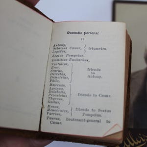 Peut inclure: Un livre ancien ouvert, affichant le titre "Dramatis Personae". La page pr&eacute;sente une liste de personnages d'une pi&egrave;ce de th&eacute;&acirc;tre. Un portrait encadr&eacute; d'un homme barbu est visible en arri&egrave;re-plan.