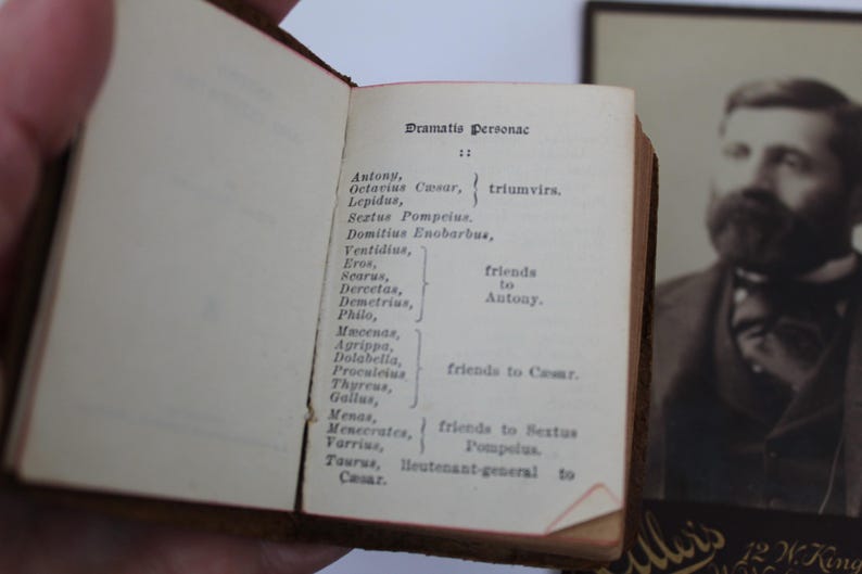 Peut inclure: Un livre ancien ouvert, affichant le titre "Dramatis Personae". La page pr&eacute;sente une liste de personnages d'une pi&egrave;ce de th&eacute;&acirc;tre. Un portrait encadr&eacute; d'un homme barbu est visible en arri&egrave;re-plan.
