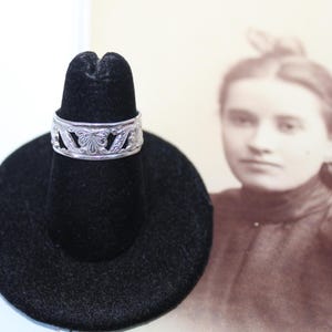 Op de afbeelding: Een zilveren ring met een ingewikkeld bloemmotief wordt gepresenteerd op een zwarte ringhouder. De ring heeft een herhalend patroon van gestileerde bladeren en bloemen. De achtergrond bevat een vintage sepia-foto van een vrouw.