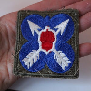 May include: Embroidered patch featuring a blue quatrefoil design with white arrows and a red acorn in the center. The patch has a green border and is likely a military or historical item.