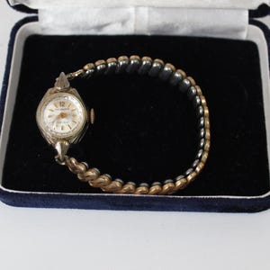 May include: A vintage wristwatch with a round silver-toned dial featuring the text "Delbana" and "17 Jewels" with gold-toned hands and hour markers. The watch has a two-tone flexible expansion band made of alternating gold and silver-toned links.