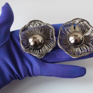 May include: A pair of silver tone earrings with a flower design. The earrings have a round, silver tone center piece.