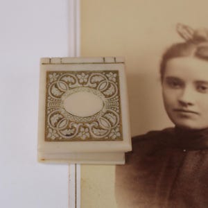 May include: Ivory-colored, rectangular box with a hinged lid. The lid features an oval center surrounded by ornate gold-colored floral and scrollwork designs. The box is positioned in front of a sepia-toned photograph of a woman.
