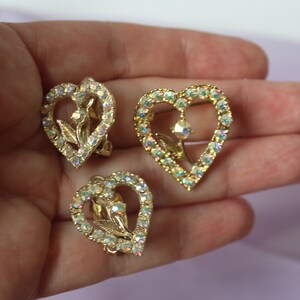 May include: A pair of gold tone heart shaped earrings with clear iridescent rhinestones. The earrings feature a floral design in the centre of the heart.