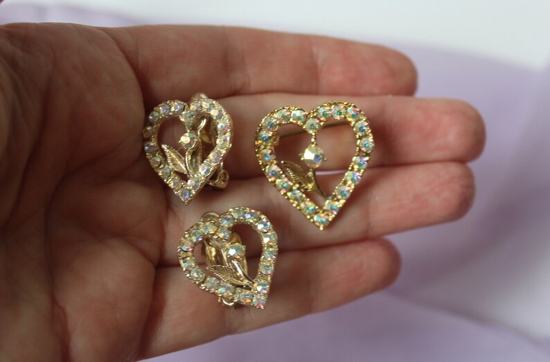 May include: A pair of gold tone heart shaped earrings with clear iridescent rhinestones. The earrings feature a floral design in the centre of the heart.