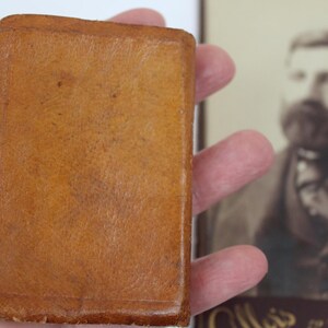 Peut inclure: Un petit livre rectangulaire ancien, reli&eacute; en cuir brun clair et chaud. La couverture du livre montre des signes d'usure, avec une surface textur&eacute;e. Un portrait en s&eacute;pia est en arri&egrave;re-plan.