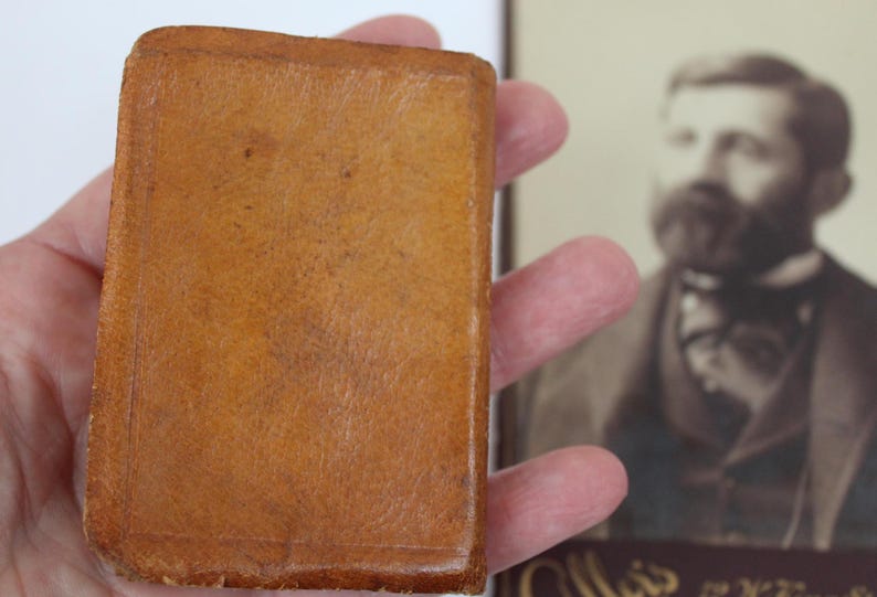 Peut inclure: Un petit livre rectangulaire ancien, reli&eacute; en cuir brun clair et chaud. La couverture du livre montre des signes d'usure, avec une surface textur&eacute;e. Un portrait en s&eacute;pia est en arri&egrave;re-plan.