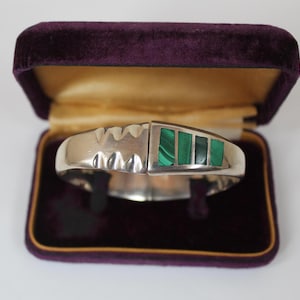 May include: A silver hinged bracelet with a malachite inlay. The bracelet is displayed open within a vintage purple velvet jewellery box with a gold interior. The malachite is green.